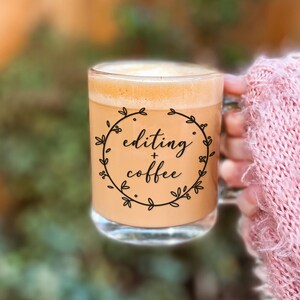 May include: A clear glass mug filled with a light brown coffee beverage. The front of the mug features a black circular wreath design with the text "editing + coffee" written in a cursive script font in the center of the wreath.