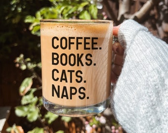 May include: A clear glass mug with a brown liquid inside. The mug has black text that reads "COFFEE. BOOKS. CATS. NAPS."
