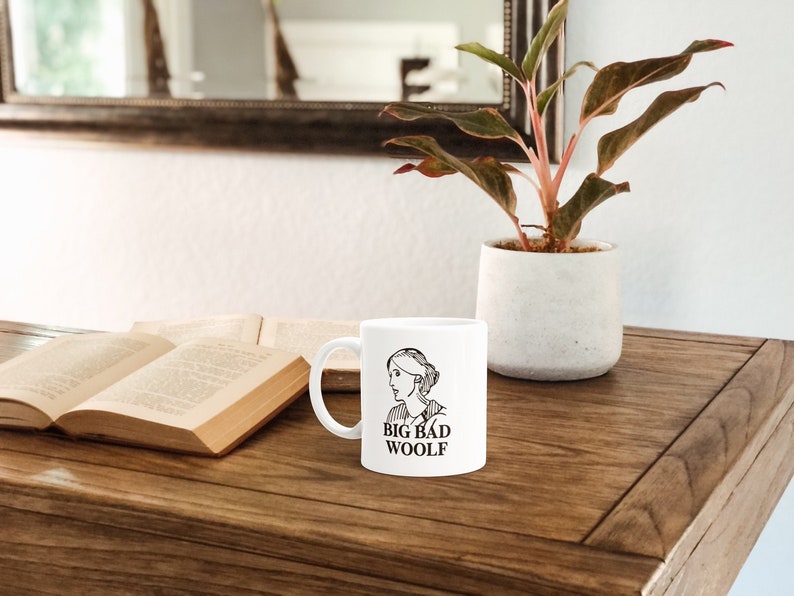 May include: White ceramic mug with a black line drawing of a woman's face and the text "BIG BAD WOOLF" on the front. The mug is sitting on a wooden table next to a book and a potted plant.