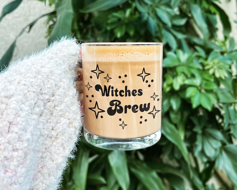 May include: A clear glass mug filled with a light brown coffee beverage, featuring black text that reads Witches Brew surrounded by various star and dot graphics printed on the front surface of the glass container.