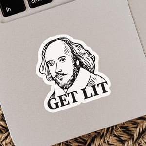May include: A white sticker featuring a black and white illustration of William Shakespeare's portrait. The sticker has the words "GET LIT" in bold, black letters below the portrait. The sticker is placed on a silver laptop.