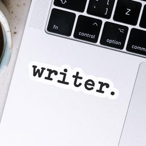 May include: A white sticker with the word "writer." in a bold, black, typewriter-style font. The sticker is placed on the silver surface of a laptop, near the keyboard.