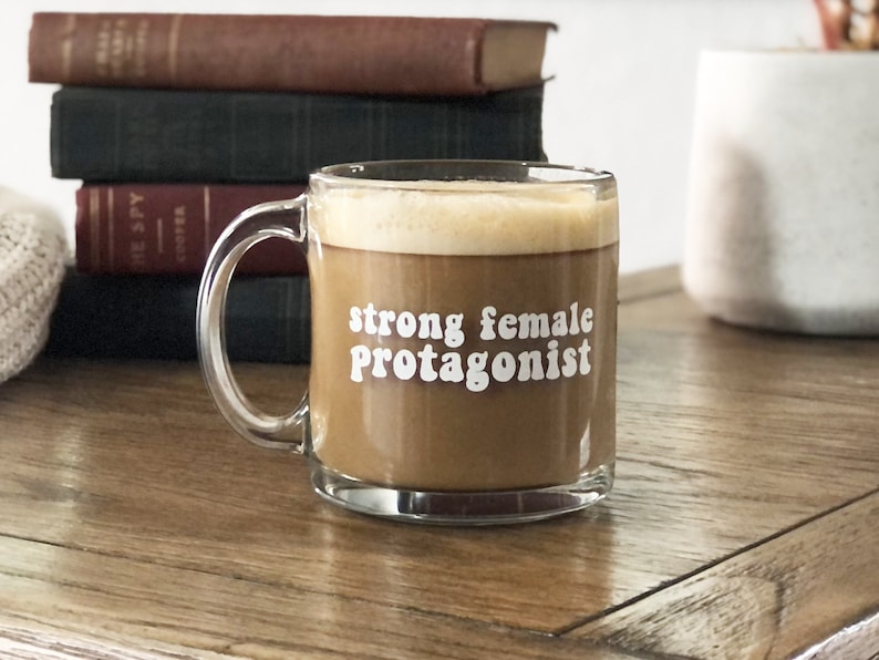 May include: A clear glass coffee mug with a brown coffee drink inside. The mug has the text "strong female protagonist" printed on it in white.