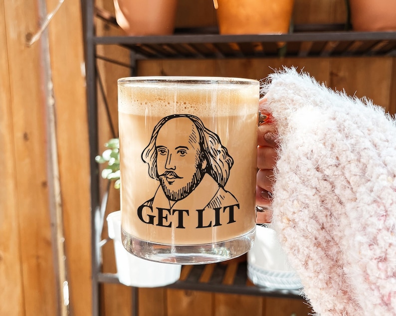 K&ouml;nnte beinhalten: Eine Glas-Tasse mit einer braunen Fl&uuml;ssigkeit darin. Die Tasse hat eine schwarz-wei&szlig;e Illustration von William Shakespeare und den Text "GET LIT" darauf gedruckt.