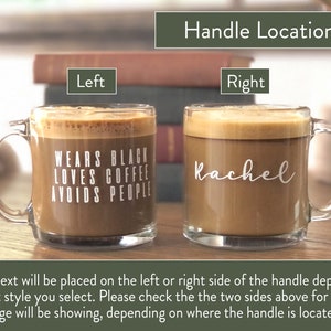 May include: Two clear glass coffee mugs with handles. The mug on the left has the text "WEARS BLACK LOVES COFFEE AVOIDS PEOPLE" printed on it. The mug on the right has the name "Rachel" printed on it. The mugs are on a wooden surface.