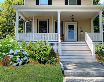 May include: A white two-story house with a front porch and steps leading to the front door. The house has a yellow exterior with white trim and black windows. The porch has a white railing and a white latticework underneath. The steps are made of wood and have a white railing. The house is surrounded by green grass and bushes.