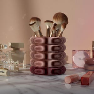 May include: A pink and burgundy makeup brush holder with a ribbed design. The holder is filled with makeup brushes and is sitting on a marble surface. There are also several makeup products on the surface, including a bottle of perfume, a compact, and a lip gloss.