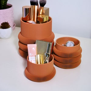 May include: Three terracotta-coloured cylindrical containers with a ribbed design. The largest container holds makeup brushes and lipsticks. The middle container holds a small bottle of lotion and a gold compact. The smallest container holds a watch.