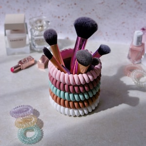 May include: A set of makeup brushes organised in a colourful hair scrunchie holder. The holder is made of multiple stacked scrunchies in pink, brown, green and white.