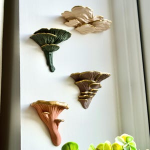 May include: Four decorative mushroom wall art pieces in shades of green, brown, and peach, with gold trim. The art is mounted on a white wall. The mushroom sculptures are designed to bring a touch of nature indoors, creating a natural aesthetic.