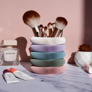 May include: A pastel-coloured makeup brush holder with a ribbed design, holding various makeup brushes. The holder is made of stacked rings in white, lilac, blue, green, and pink. Other beauty products are visible in the background.