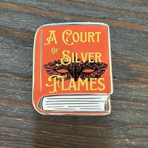 May include: Orange enamel pin in the shape of a book with the title "A Court of Silver Flames" in gold lettering. A black masquerade mask design is centered on the book cover. The pin has a silver border and white pages.