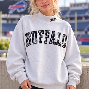 May include: A light gray mock neck sweatshirt featuring the word BUFFALO in black and white gingham block letters across the chest and the word Bills embroidered in black script on the center of the collar neckline.
