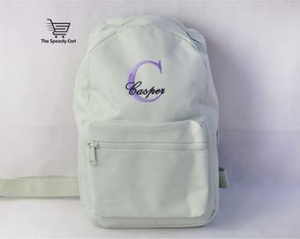 May include: A light mint green backpack featuring a large embroidered purple letter C and the name Casper in black script. The bag includes a front zippered pocket and matching mint green straps. The logo The Speedy Cart is visible in the corner.