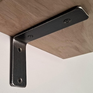 May include: A close-up of a wooden shelf supported by a black metal L-shaped bracket. The shelf is made of light-colored wood, and the bracket is attached with screws. The bracket is mounted on a white wall.