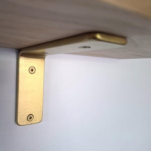 Pode incluir: Um suporte de prateleira de metal em forma de L com acabamento em dourado escovado. O suporte está fixado a uma parede branca e à parte inferior de uma prateleira de madeira clara usando quatro parafusos visíveis com fenda cruzada.