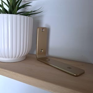 Pode incluir: Um suporte de prateleira de metal em forma de L na cor dourada com quatro orifícios para parafusos pré-perfurados, posicionado numa prateleira de madeira clara ao lado de um vaso de cerâmica branco com estrias verticais e uma suculenta verde.
