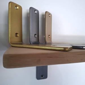 Pode incluir: Três suportes de prateleira de metal em forma de L com acabamentos em dourado, prateado e bronze, cada um com dois orifícios circulares para parafusos no braço vertical e dois no horizontal, montados para apoiar uma prateleira de madeira clara.