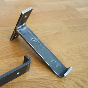 Pode incluir: Dois suportes de prateleira de metal, um é uma forma simples de L e o outro é um design mais elaborado com uma borda curva. Ambos são de metal escuro e têm um aspecto áspero e industrial.