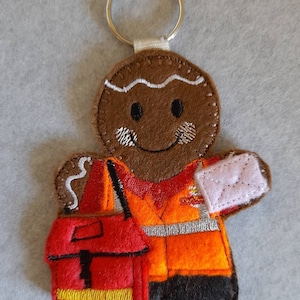 May include: A gingerbread man wearing a brown, orange and black postal worker uniform. He is holding a pink envelope and a red bag with a yellow strap. The gingerbread man has a white icing smile and black eyes.