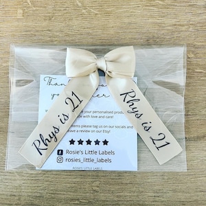 May include: A beige satin ribbon bow with the text "Rhys is 21" printed in black. The bow is packaged in a clear plastic bag with a small card that reads "Rosie's Little Labels". The background is a light brown wooden surface.