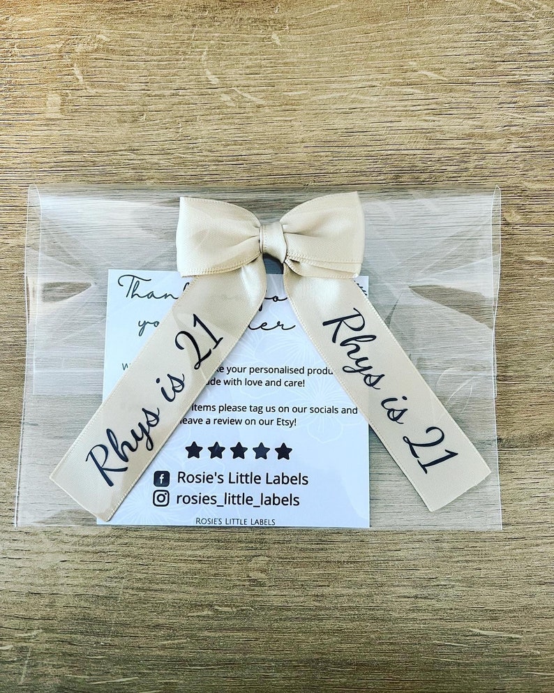 May include: A beige satin ribbon bow with the text "Rhys is 21" printed in black. The bow is packaged in a clear plastic bag with a small card that reads "Rosie's Little Labels". The background is a light brown wooden surface.