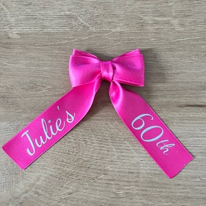 May include: A vibrant pink satin ribbon bow, featuring the text "Julie's 60th" in white. The bow is centrally positioned, with the ribbon tails spreading out. The backdrop is a light wood grain.