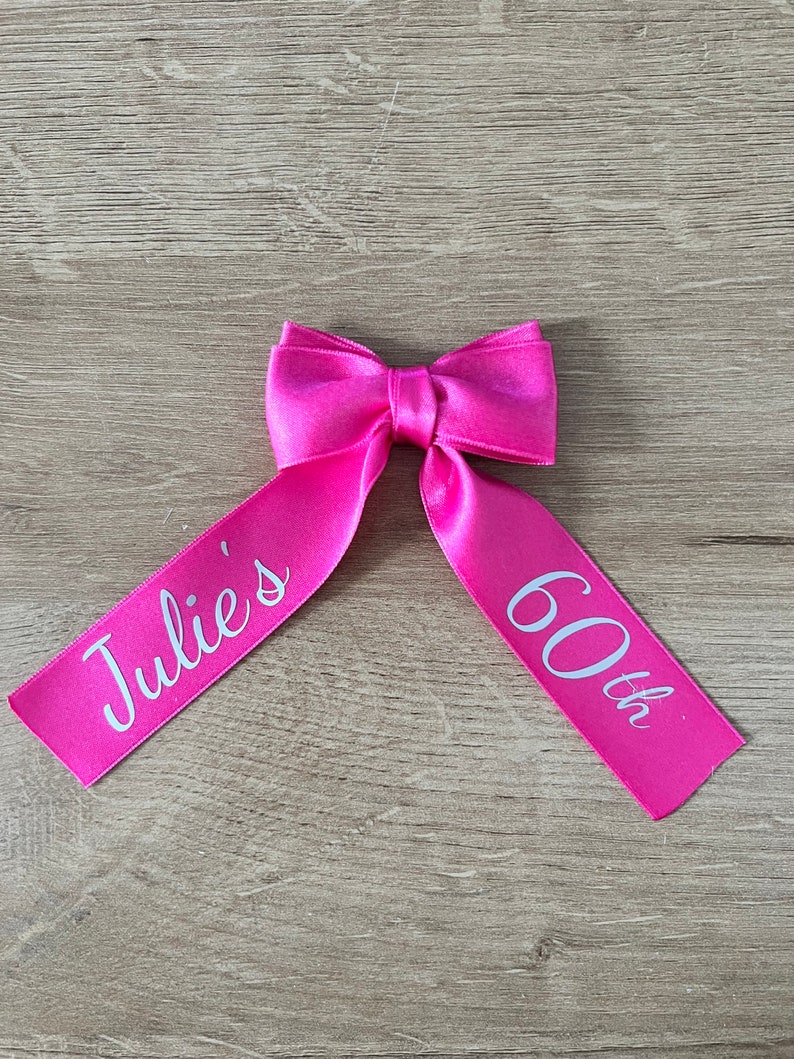 May include: A vibrant pink satin ribbon bow, featuring the text "Julie's 60th" in white. The bow is centrally positioned, with the ribbon tails spreading out. The backdrop is a light wood grain.