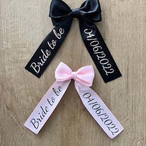May include: Two satin bows, one black and one pink, with white text. The black bow reads "Bride to be" and "04/06/2022". The pink bow reads the same. The bows are on a wooden surface.