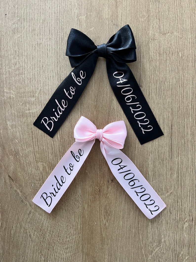 May include: Two satin bows, one black and one pink, with white text. The black bow reads "Bride to be" and "04/06/2022". The pink bow reads the same. The bows are on a wooden surface.