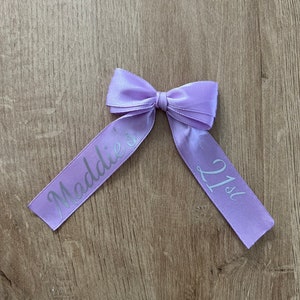 May include: A lilac satin ribbon bow with the text "Maddie's 21st" printed in silver. The bow is tied in the centre and rests on a wooden surface. The ribbon is a pale purple colour.