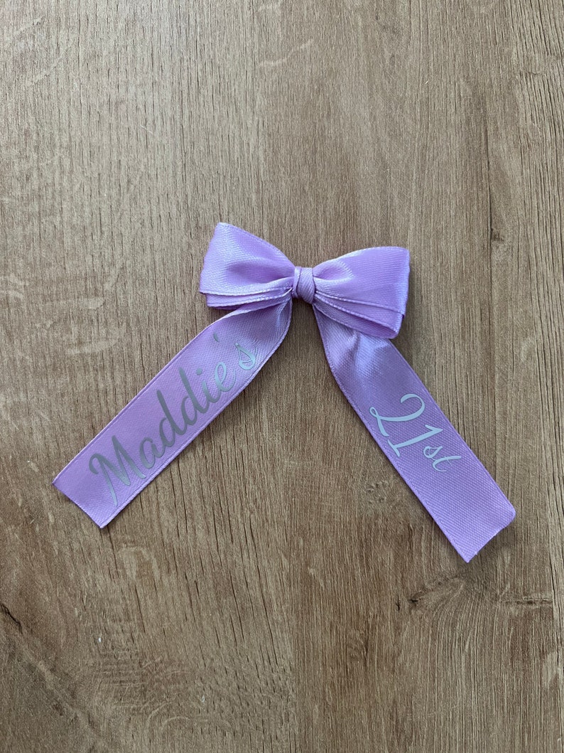 May include: A lilac satin ribbon bow with the text "Maddie's 21st" printed in silver. The bow is tied in the centre and rests on a wooden surface. The ribbon is a pale purple colour.