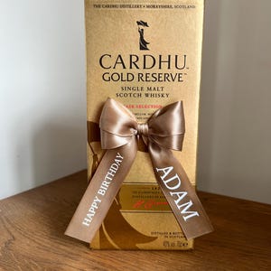 May include: A gold-coloured box of Cardhu Gold Reserve Single Malt Scotch Whisky, with a brown satin ribbon tied in a bow. The ribbon has the words "Happy Birthday" and "Adam" printed on it in white. The box has text that reads "The Cardhu Distillery, Morayshire, Scotland."