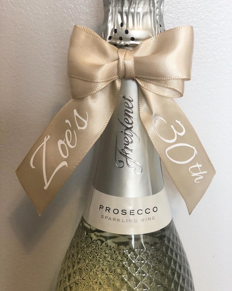 May include: A bottle of Prosecco sparkling wine with a champagne-colored ribbon tied in a bow. The ribbon is printed with the words "Zoe's 30th" in a cursive font. The bottle has a silver foil top.
