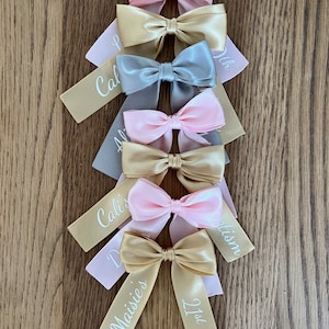 May include: An assortment of satin bows in pink, gold, and grey, displayed on a wooden surface. Each bow features a ribbon tail with white personalised text, including names and the number "21st". The bows are tied in a classic bow style.