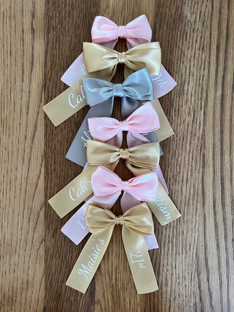 May include: An assortment of satin bows in pink, gold, and grey, displayed on a wooden surface. Each bow features a ribbon tail with white personalised text, including names and the number "21st". The bows are tied in a classic bow style.