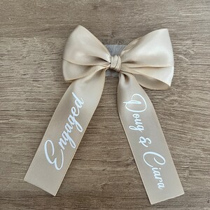 May include: A beige satin bow with the word "Engaged" printed on one tail and "Doug & Ciara" on the other in white script. The bow is tied and ready to use.