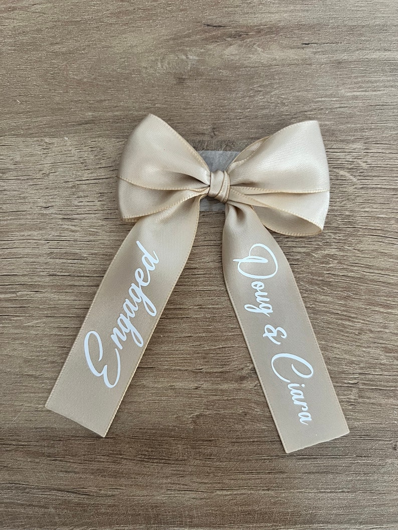 May include: A beige satin bow with the word "Engaged" printed on one tail and "Doug & Ciara" on the other in white script. The bow is tied and ready to use.