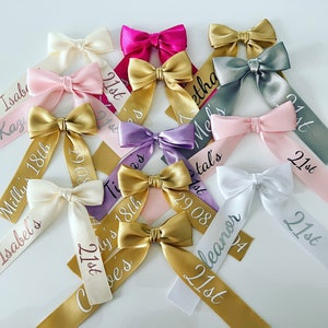 May include: A collection of satin ribbon bows in various colors, including pink, gold, and white. Each bow has a personalized ribbon tail with names, dates, and the text "21st" or "18th" in a script font. These are likely for a birthday celebration.