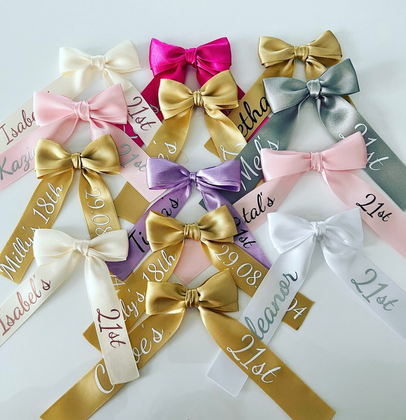 May include: A collection of satin ribbon bows in various colors, including pink, gold, and white. Each bow has a personalized ribbon tail with names, dates, and the text "21st" or "18th" in a script font. These are likely for a birthday celebration.
