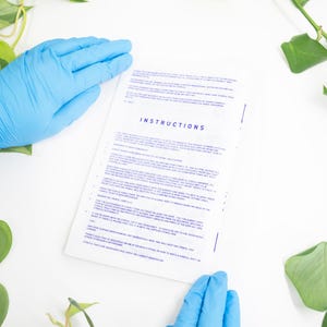 May include: A white sheet of paper with the word "INSTRUCTIONS" in blue text is held by two hands wearing blue gloves. Green leafy plants surround the paper on a white surface. The instructions appear to be for a stencil.