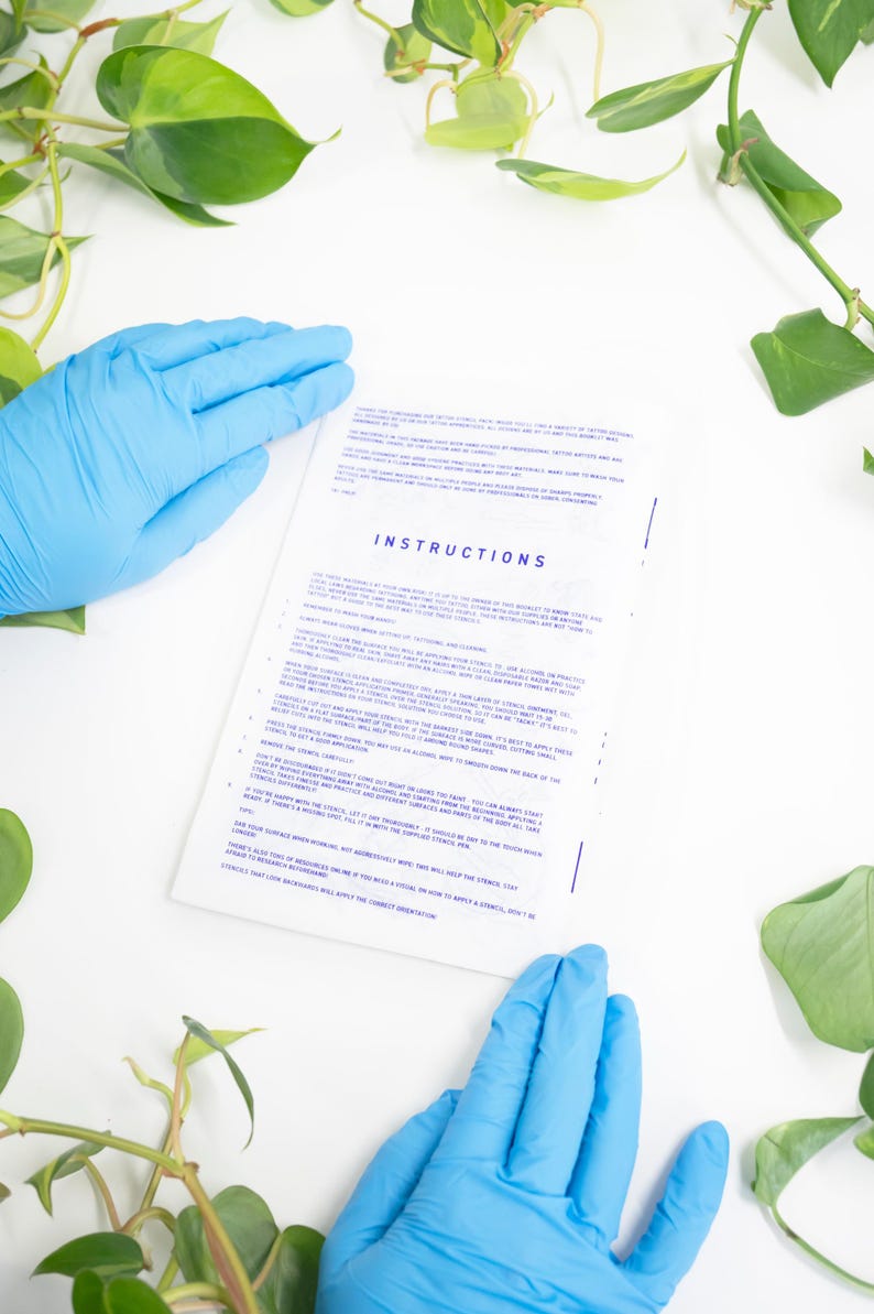 May include: A white sheet of paper with the word "INSTRUCTIONS" in blue text is held by two hands wearing blue gloves. Green leafy plants surround the paper on a white surface. The instructions appear to be for a stencil.