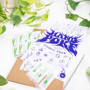 May include: A white stencil pack with blue graphics and text, including the words "Stencil Pack" and "18+". The pack is accompanied by small packets and a brown envelope, with a silver tool. Green plants are in the background.