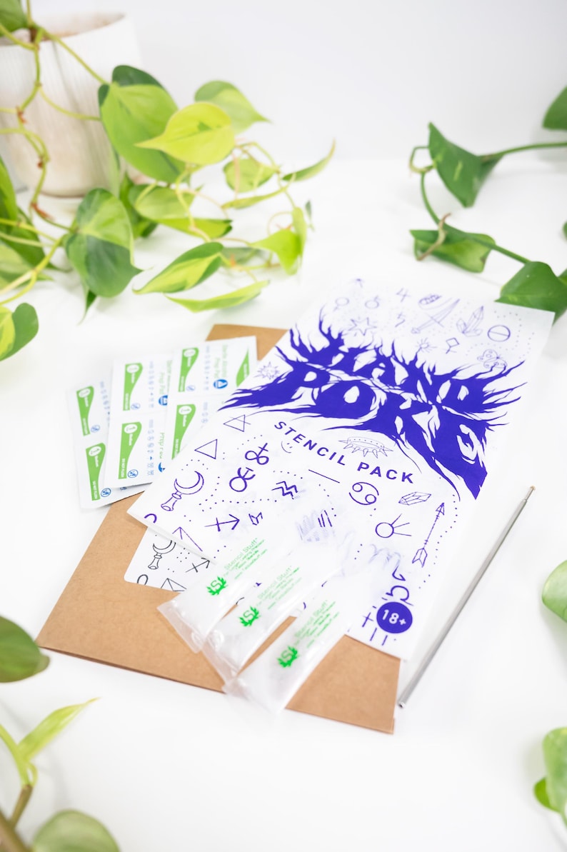 May include: A white stencil pack with blue graphics and text, including the words "Stencil Pack" and "18+". The pack is accompanied by small packets and a brown envelope, with a silver tool. Green plants are in the background.