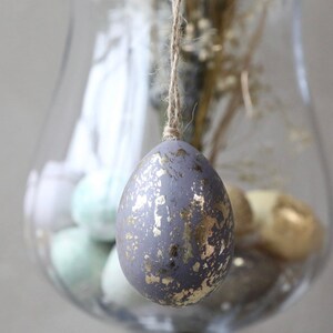 May include: A grey Easter egg with gold flecks hanging from a string in a glass vase. The egg is surrounded by other Easter eggs in various colours.