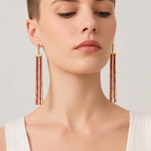 Può includere: Un paio di lunghi orecchini pendenti con un perno dorato e una barra orizzontale da cui pendono diversi fili verticali di piccole perline alternate rosse e dorate, creando un effetto a frange stratificato che arriva fino alla linea della mascella.