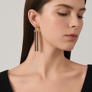 Può includere: Un orecchino pendente color oro caratterizzato da un design a barra verticale con file alternate di piccole perline dorate e nere. L'orecchino pende da un perno a sfera dorata, creando un effetto a frangia lineare a più fili.