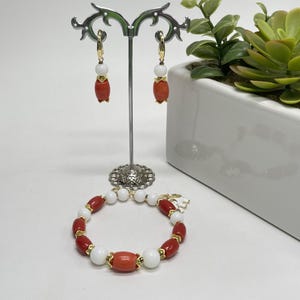 Può includere: Un set di gioielli con bracciale e orecchini. Il bracciale e gli orecchini presentano una combinazione di perline di corallo bianco e rosso con dettagli dorati. Gli orecchini sono esposti su un supporto argentato. Sullo sfondo una fioriera bianca con vegetazione.