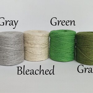 Op de afbeelding: Vier strengen touw in verschillende kleuren: grijs, gebleekt, groen en grasgroen. Elke streng is gelabeld met de kleurnaam. Het touw is strak opgerold, geschikt voor knutselen of verpakken.