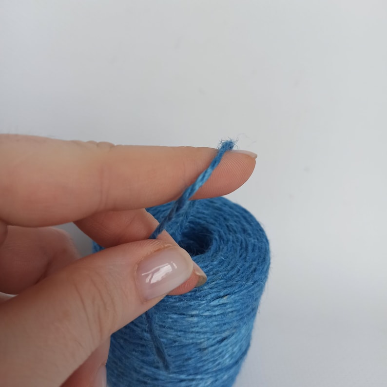 Op de afbeelding: Een close-up van een blauwe kluwen touw met een draad die door een vinger wordt vastgehouden. Het touw is een levendige blauwe tint en de kluwen lijkt te zijn gemaakt van strak gewikkelde vezels. De achtergrond is effen wit.